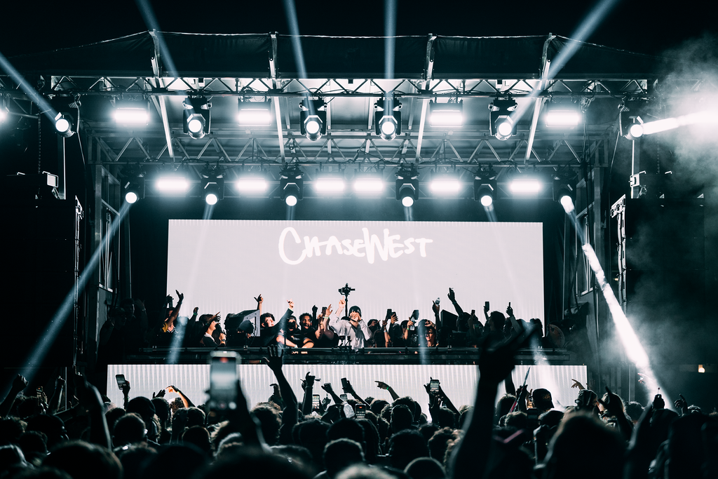 Crowd at a nighttime concert facing a large illuminated stage with a digital screen showing 'ChaseNest' and hands raised in the air.